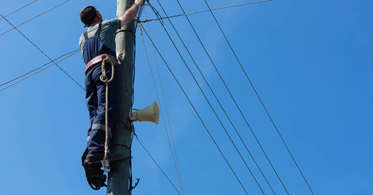 An electrician wearing bib overalls is hooked to a wooden electrical pole. They are reaching up with one arm.