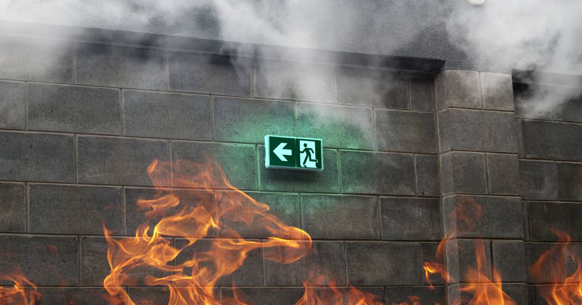 In a room with flames and smoke, a green emergency exit sign on a stone wall shows an arrow pointing to the left.