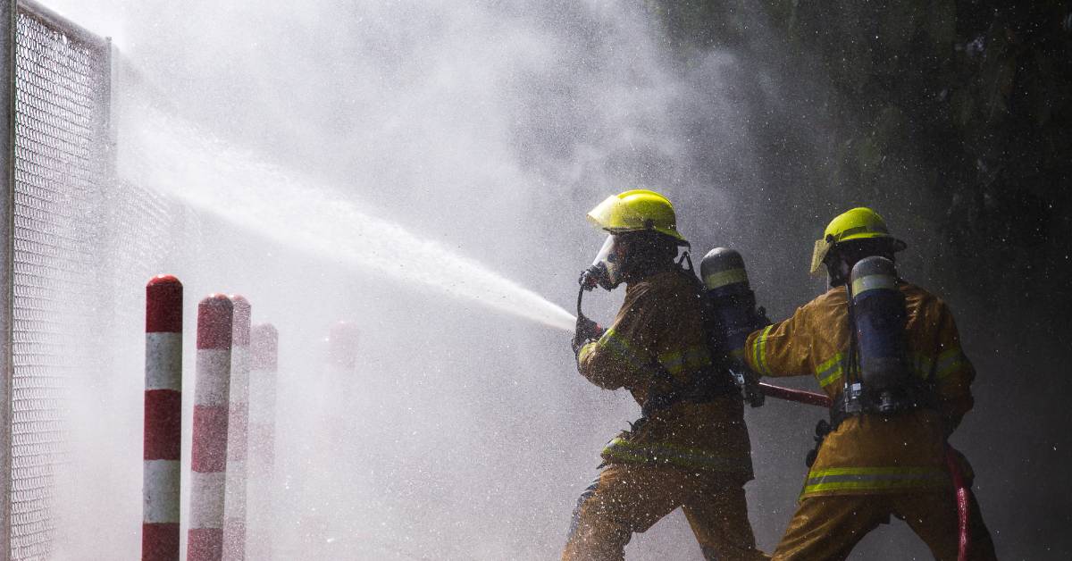 Two firefighters standing behind a gate and holding a hose. The hose is spraying water through the gate.