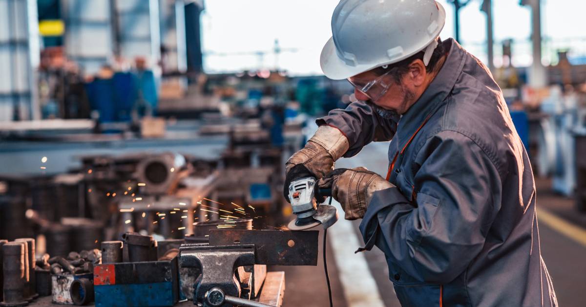 A man using a tool to work on some metal. He's wearing gloves, a helmet, eye protection, and other protective clothing.