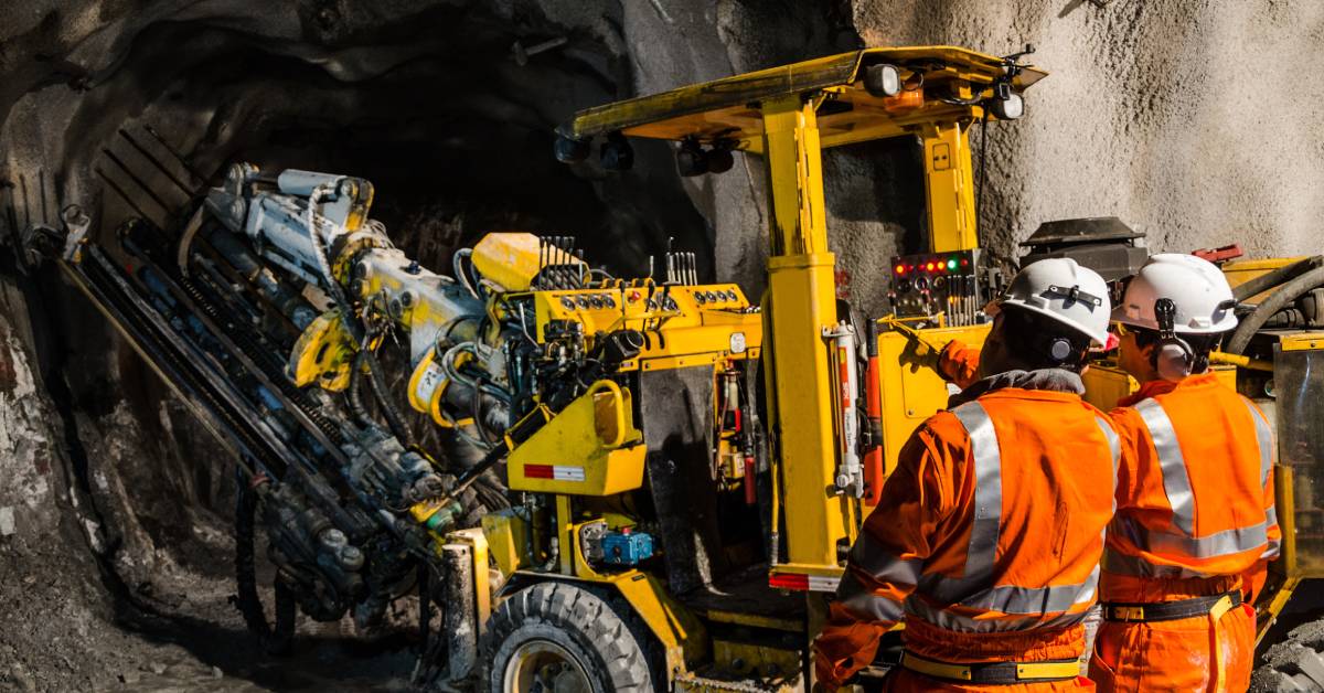 Two people inside of a mine, with one of them pointing at something. They're wearing orange protective clothing and hard hats.