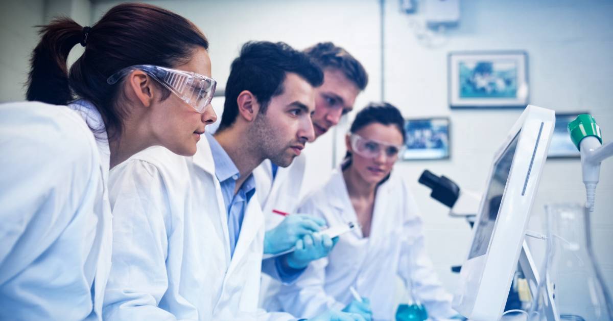 Multiple people inside a lab looking at a computer screen. They are all wearing lab coats and two are wearing eyeglasses.