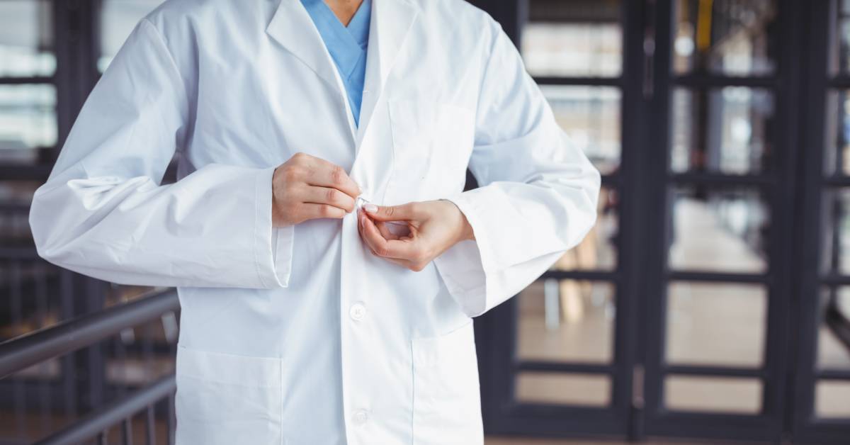 A person wearing blue scrubs buttoning up a white lab coat. The person is standing next to a dark rail.