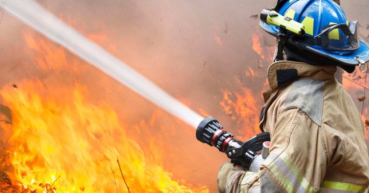 A firefighter wearing protective gear stands very close to a fire and sprays water using a red hose.