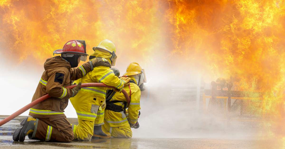 Three firefighters kneeling on the ground and holding on to a hose. The hose is spraying water at a fire.