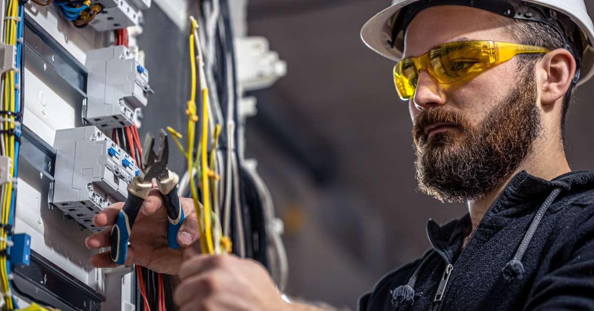 An electrician working on some electrical cables with some pliers. He's wearing a helmet, a jacket, and eyeglasses.