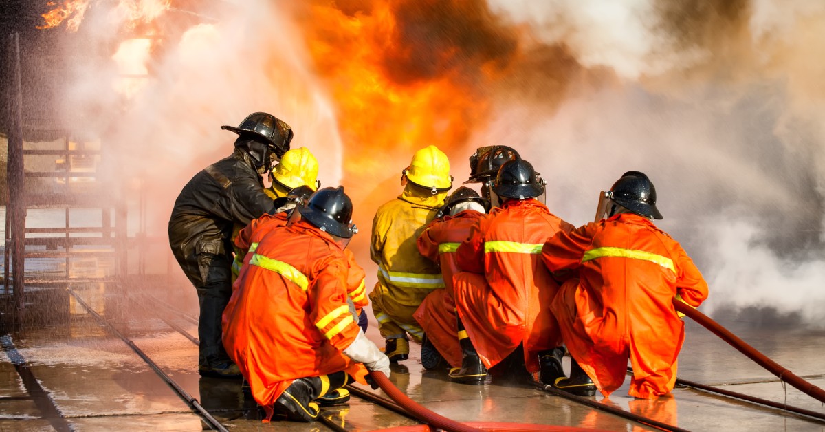 Two groups of firefighters holding hoses as they spray water at a fire. Most of the firefighters are crouched on the ground.