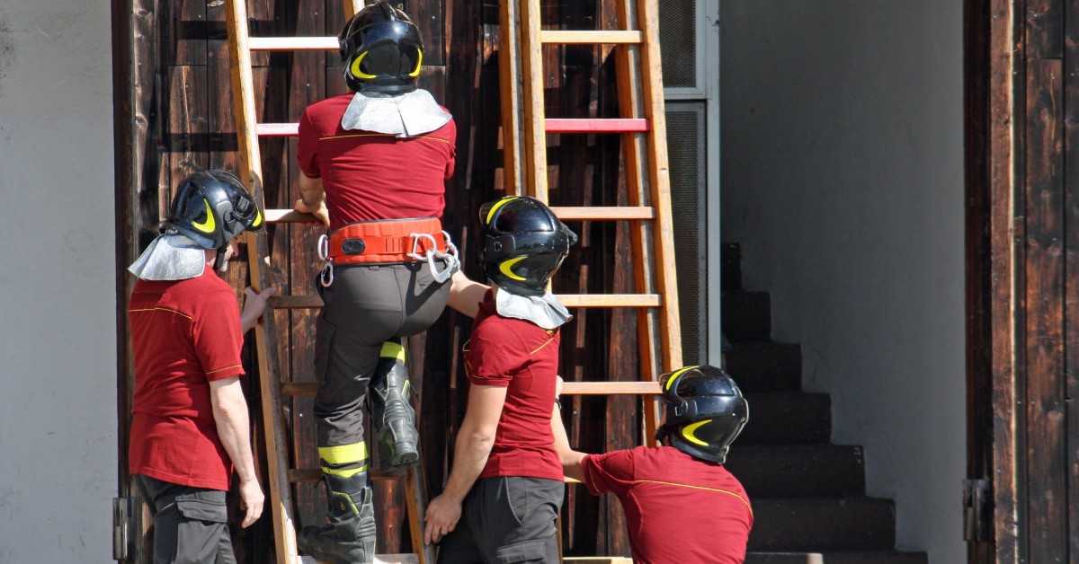 Two firefighters are holding a ladder as one firefighter is climbing it. Another firefighter is holding a separate ladder.