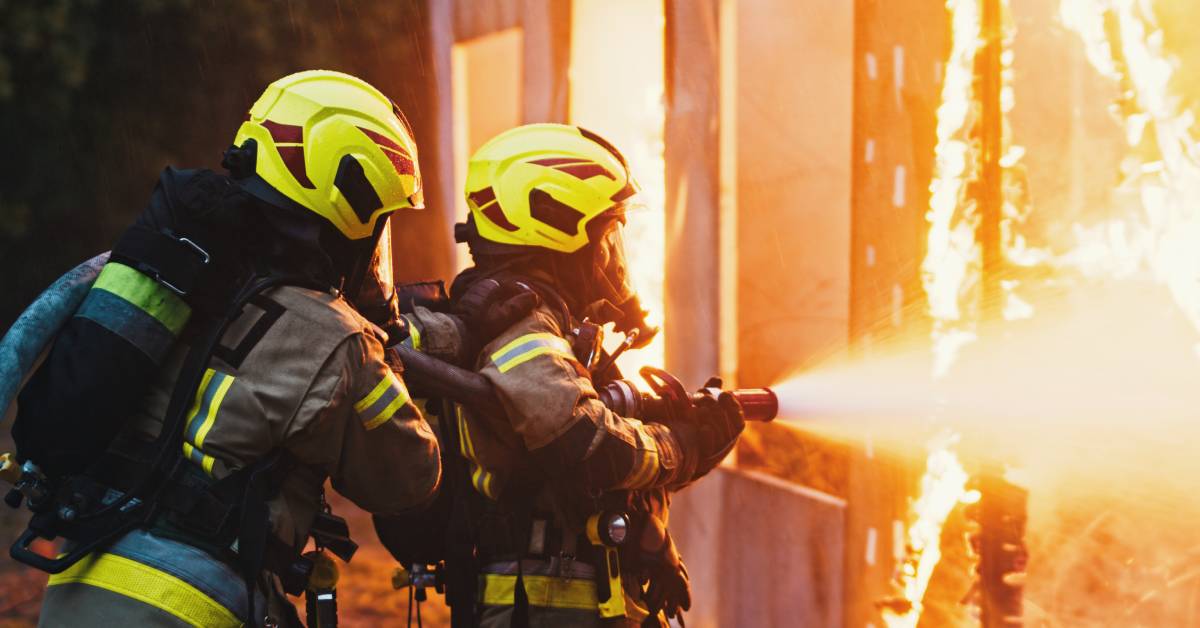 Two firefighters working close to a burning house. They are wearing firefighting gear while holding a hose and spraying the house.