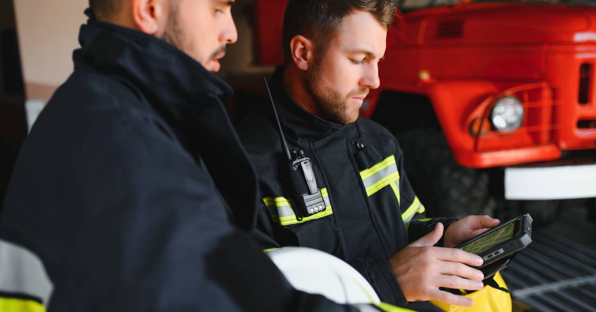 Two men wearing firefighting gear while standing together to look at a tablet. There is a red vehicle in the background.