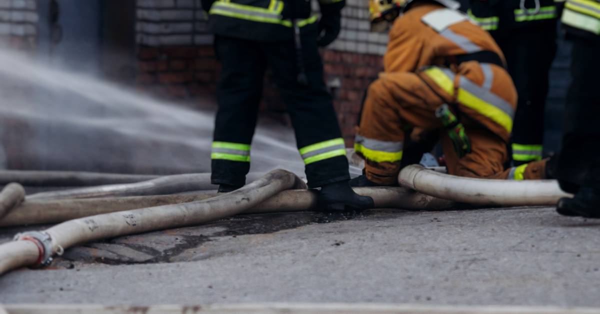 Multiple firefighters near a hose that's spraying water. One of the firefighters is crouched on the ground.