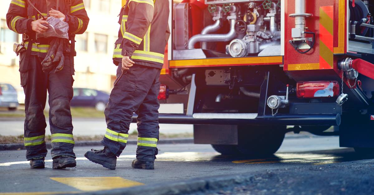 Two firefighters standing near a fire truck. The firefighters are wearing fire gear and holding fire equipment.