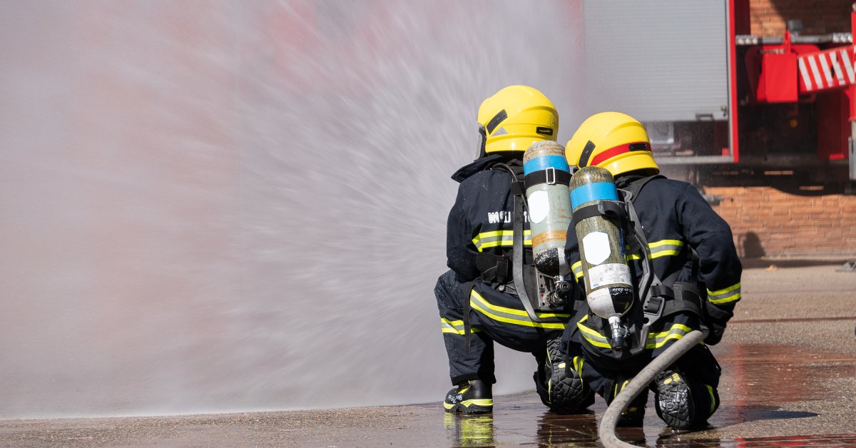 Two firefighters kneeling on the ground while holding a hose that is spraying water. There is a brick building in the background.