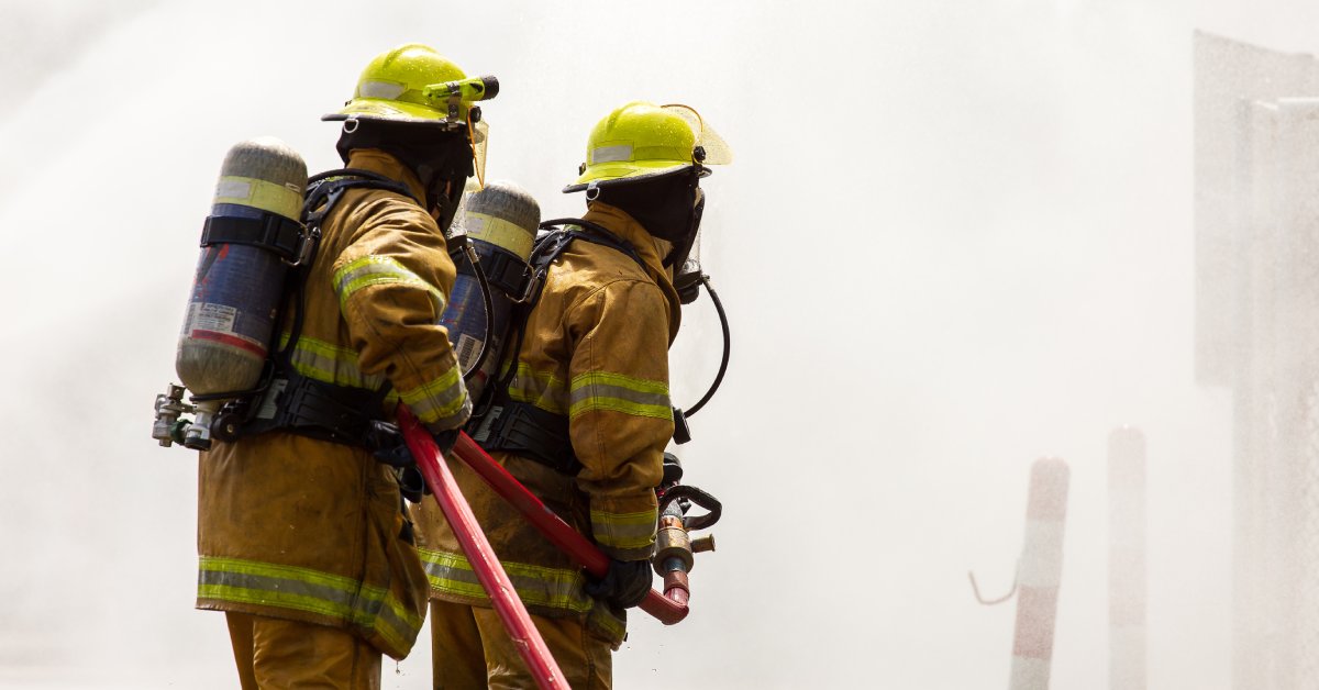 Two firefighters wearing fire-resistant clothing and yellow helmets. They are holding a hose and facing a wall of smoke.