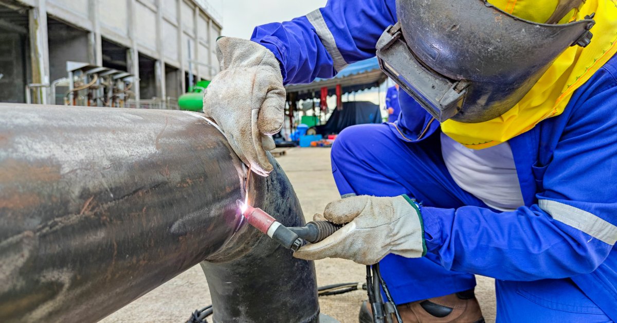 A welder working on a pipe with a welding tool. They are wearing protective clothing, gloves, and a face shield.