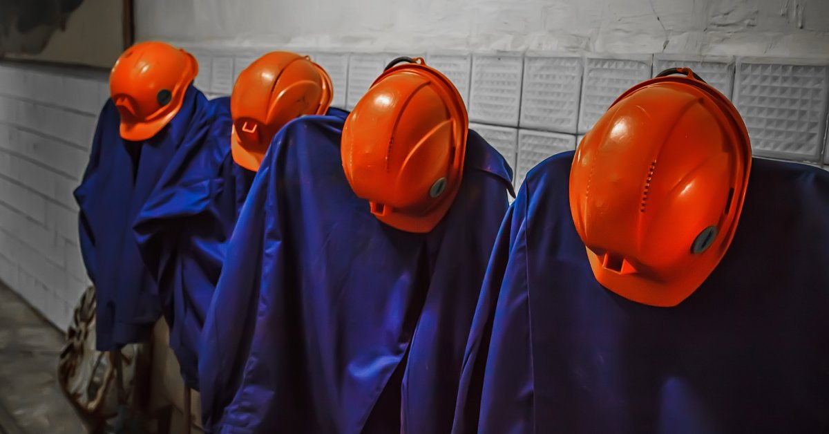 Four sets of blue coveralls and orange helmets hanging on hooks on a wall. The wall behind the gear is gray.