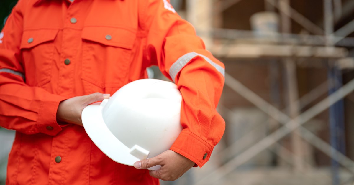 A person wearing an orange coverall uniform and holding a white helmet. There's a structure in the background.