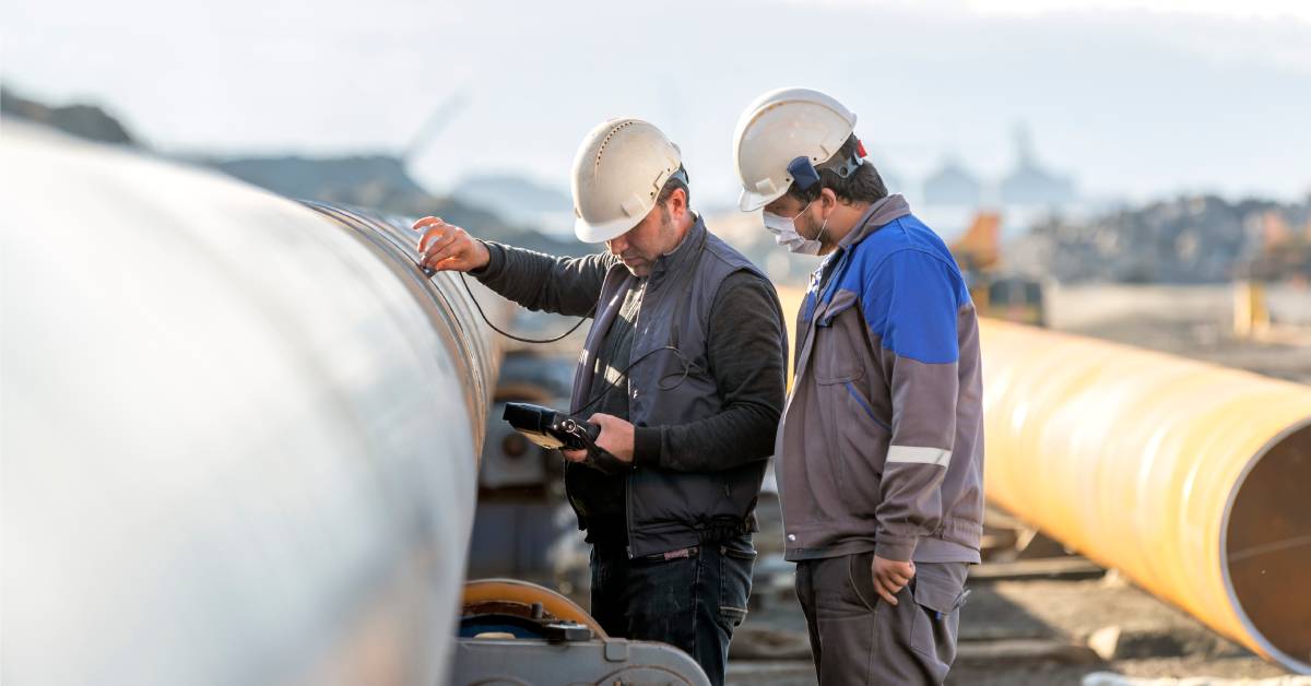 Two men wearing helmets and looking at a pipe. The men are outside, and one of them is wearing a face mask.