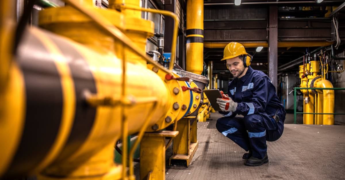 A person crouched next to a yellow pipeline inside of a building. The person is using a device and wearing a helmet.