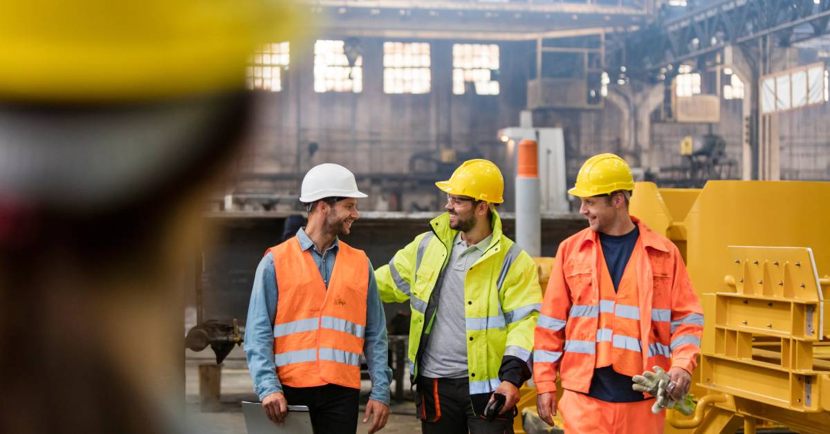 Three men wearing hard hats and yellow and orange fire-resistant clothing walk together, talking and smiling.