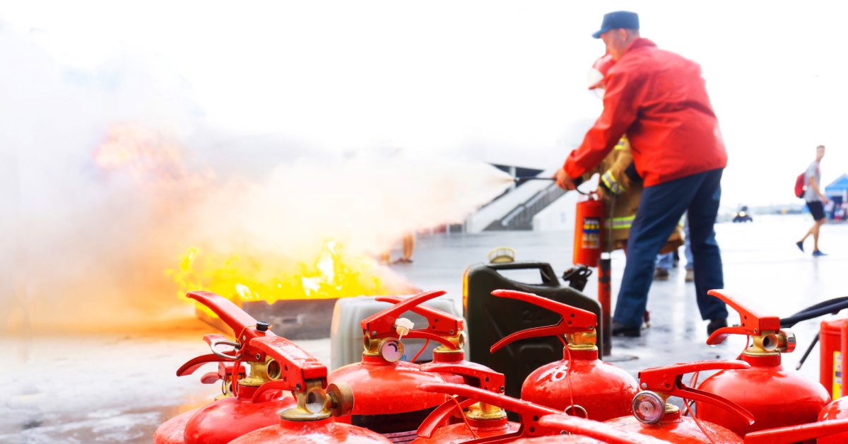 A group of red fire extinguishers sit on the ground in front of a man using one to put out a fire.