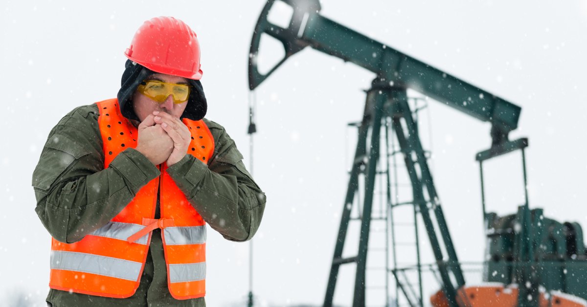 A man in an orange hard hat and vest tries to warm himself by blowing into his hands. He's standing in a snowy oil field.