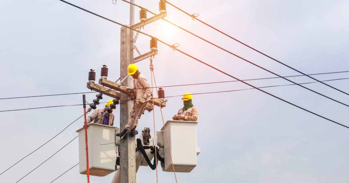 Three electrical line workers work up in the blue sky on an electrical line pole, wearing tan protective clothing.