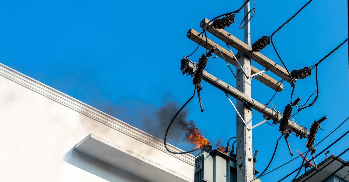 The transformer at the top of an electrical pole is on fire, spewing flames and smoke into a blue sky.