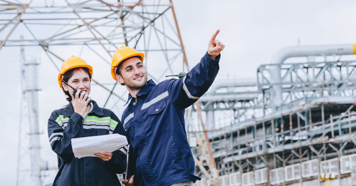 A male and female electrical worker stand together in blue FR clothing at an electrical facility