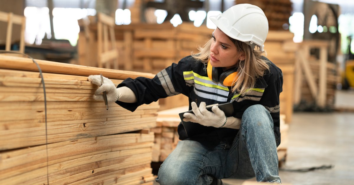 A female industrial worker in an FR shirt and white hard hat squats and inspects a pile of wooden planks.