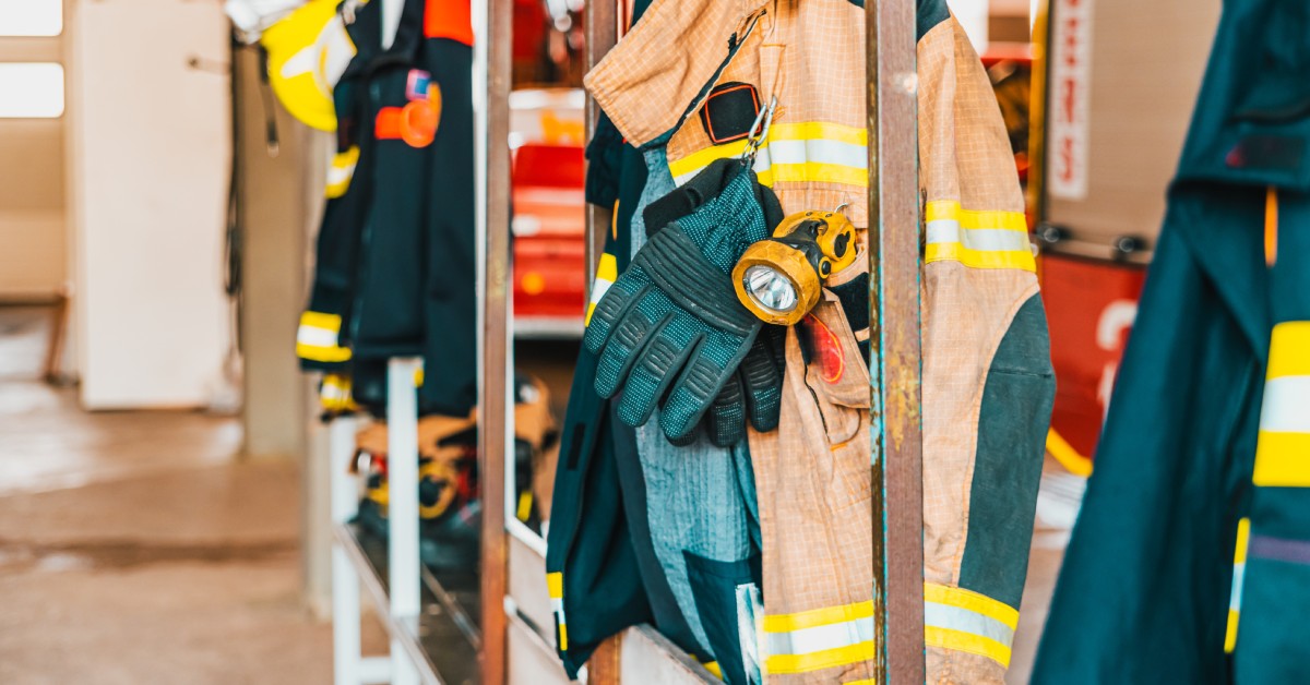 Various pieces of fire-resistant gear, including helmets, gloves, and jackets, hang on hooks in a fire station.