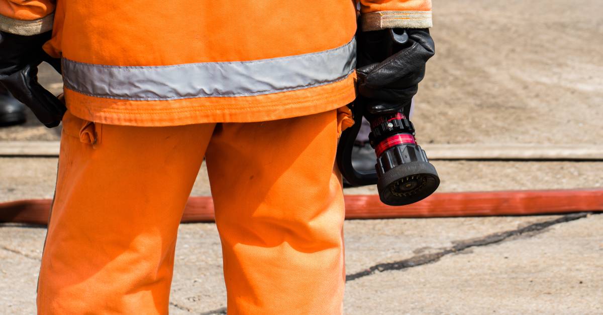 The backside of a person wearing a protective orange jacket and pants with black gloves. He holds the nozzle of a hose.