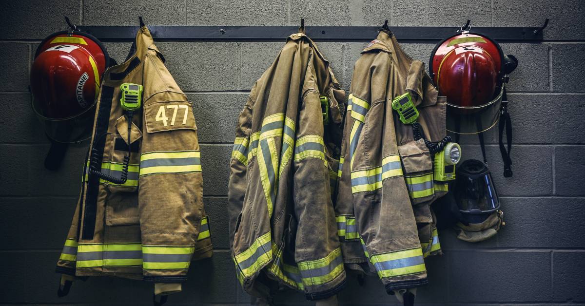 Three fire-resistant jackets and two red helmets hanging from a set of hooks attached to a concrete block wall.