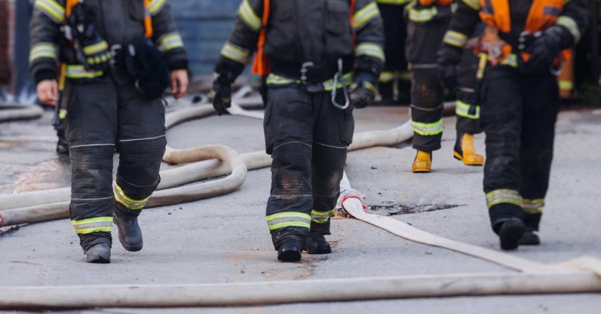 Three firefighters wearing uniforms walk around a large hose. There are other firefighters in the background.