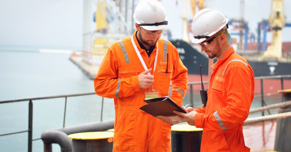 Two men wearing orange coveralls, safety glasses, and helmets. They're looking at a folder with paper in it.