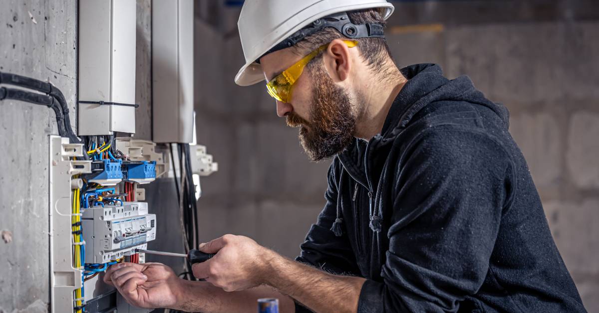 A man working on an electrical switchboard with a tool. He's wearing a hard hat, eye protection, and a sweatshirt.