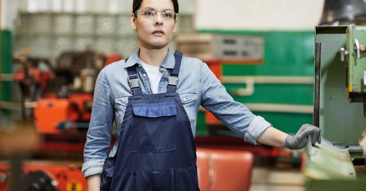 A woman wearing protective glasses, gloves, and blue bib overalls stands next to factory machinery.