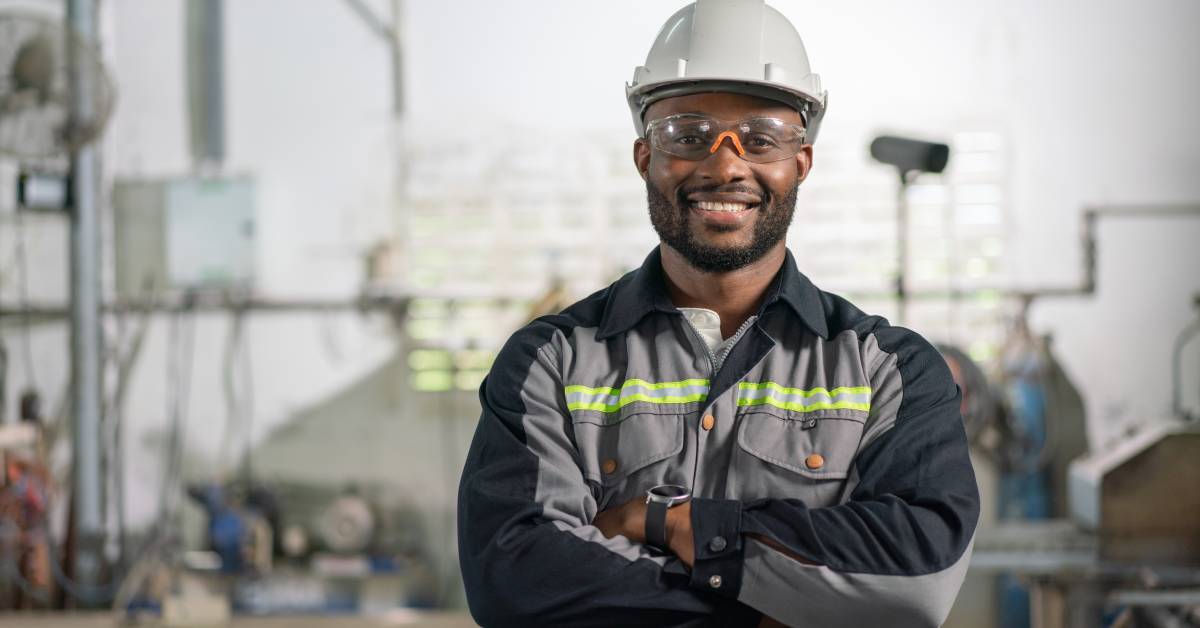 A man wearing a white hard hat, safety glasses, and a gray FR outfit stands with his arms crossed and smiles.