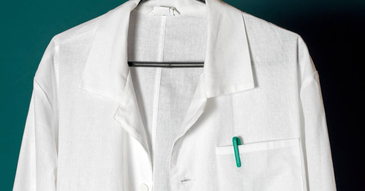 A white lab coat with white buttons hanging on a black hanger. The pocket has a blue pen inside of it.