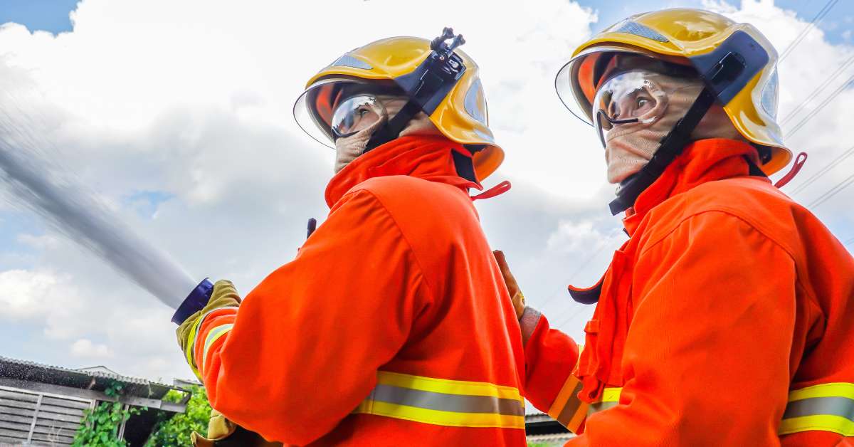 Two people wearing fire-resistant gear are both holding a hose that is spraying water. It's a sunny day.