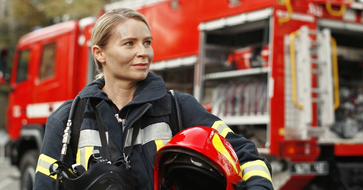 A woman wearing firefighter gear and looking off to the side. There's a fire truck behind her, and she's holding a helmet.