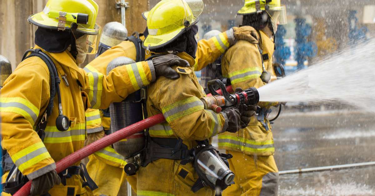 A group of firemen holding a hose that is spraying water. The firemen are wearing firefighting gear and the hose is red.