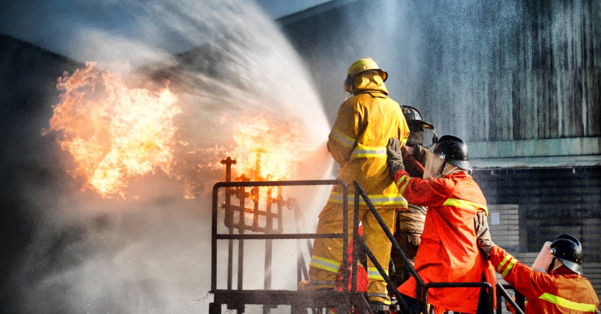 Firefighters standing on stairs and holding a hose that's spraying at a fire. They're wearing fire gear.