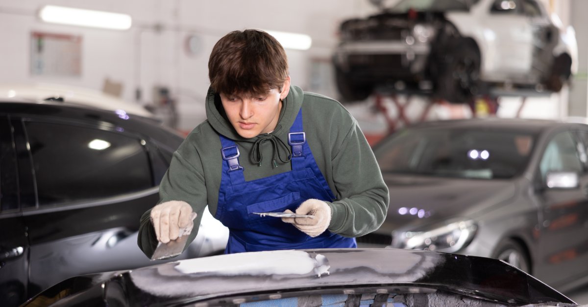 A man wearing a green hoodie, gloves, and blue FR bib overalls works on a car in a garage.